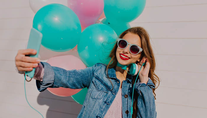 Adolescente avec des lunettes de soleil et un casque autour du coup, regardant son téléphone de loin avec des ballons gonflables rose et bleu aqua en fond